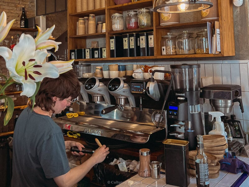 A barista preparing to brew coffee behind the bar and a wooden cabinet with coffee cups and other powdered drinks in one of the best coffee shops in Seville Spain