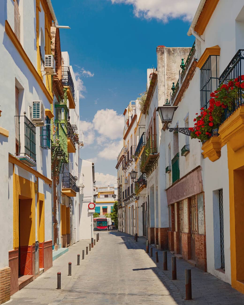 The cobbledstone street of the Barrio de Triana surrounded by lines of white buildings with accents of yellow paint and potted red flowers hanging under sunny skies to pass by during Seville bike tours