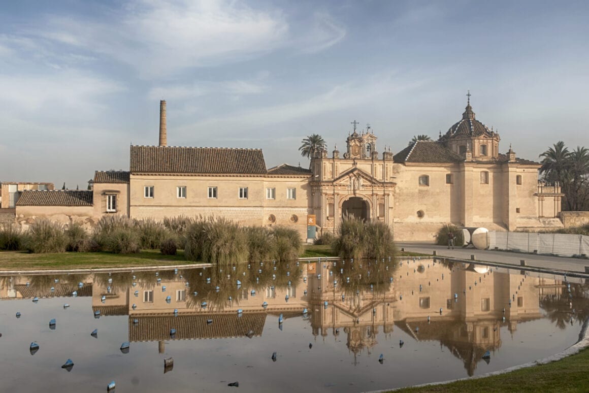 A pond in front of the Monastery of Santa Maria de las Cuevas with a road passed by people during Seville bike tours under skies with faint clouds.