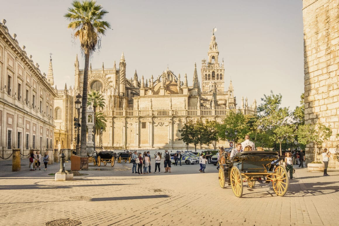 A sunny square of the Seville Cathedral filled with people walking, cars parked, and carriage with people on them. This place is a top destination for Seville bike tours