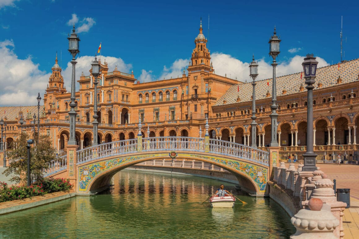 Two people rowing a boat under the bridge decorated with flower murals with the background of the Plaza de Espaรฑa, a top destination in the Seville bike tours