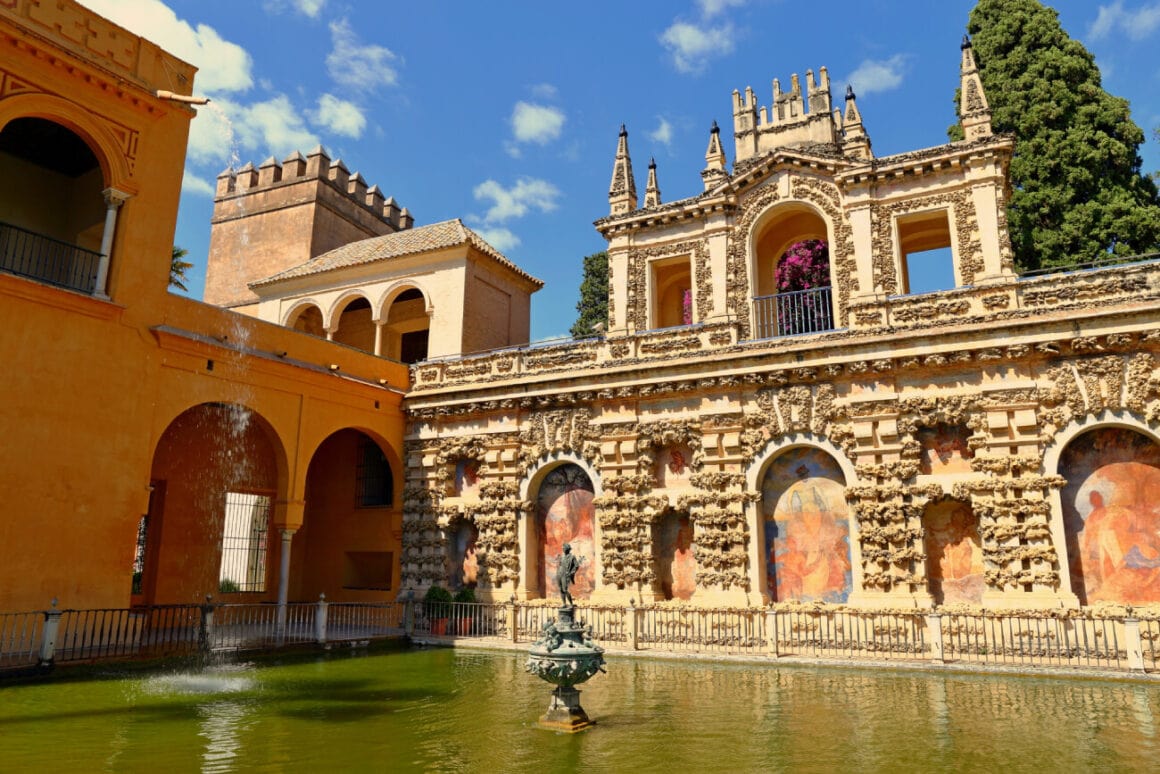 A pond with a small statue in the center of the Plaza de Espaรฑa surrounded by buildings painted in orange with a tree in the background under cloudy blue skies, a destination in Seville bike tours
