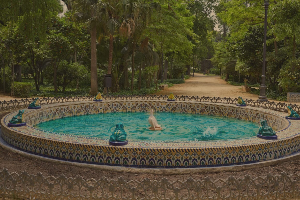 A circular fountain beaming in blue colored water while decorated by tiles with patterns in blue and yellow while small frog statues surround it. A path leading to the forest in the background can be seen that Seville bike tours usually pass by.