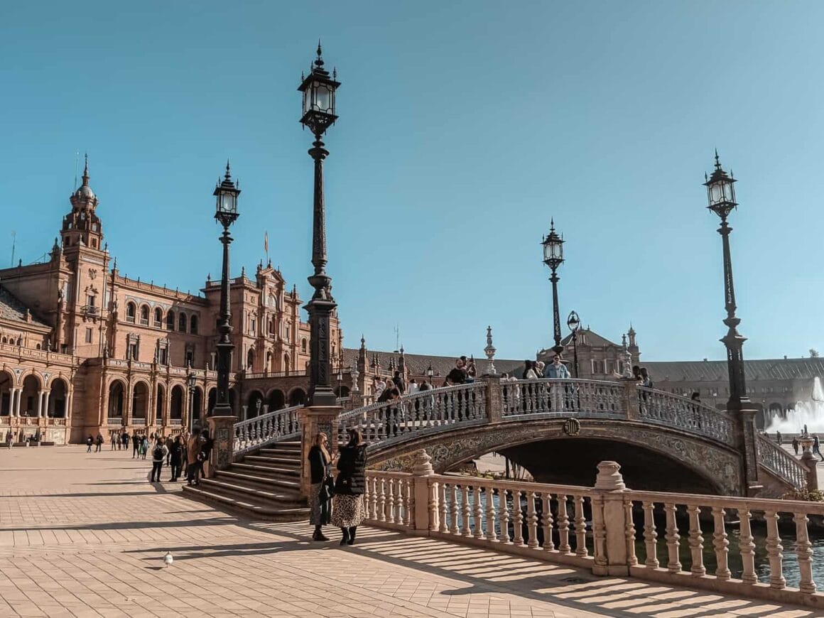 Street with a river under a bridge with people walking in front of the Plaza de Espaรฑa under clear blue skies to pass by when joining Seville bike tours