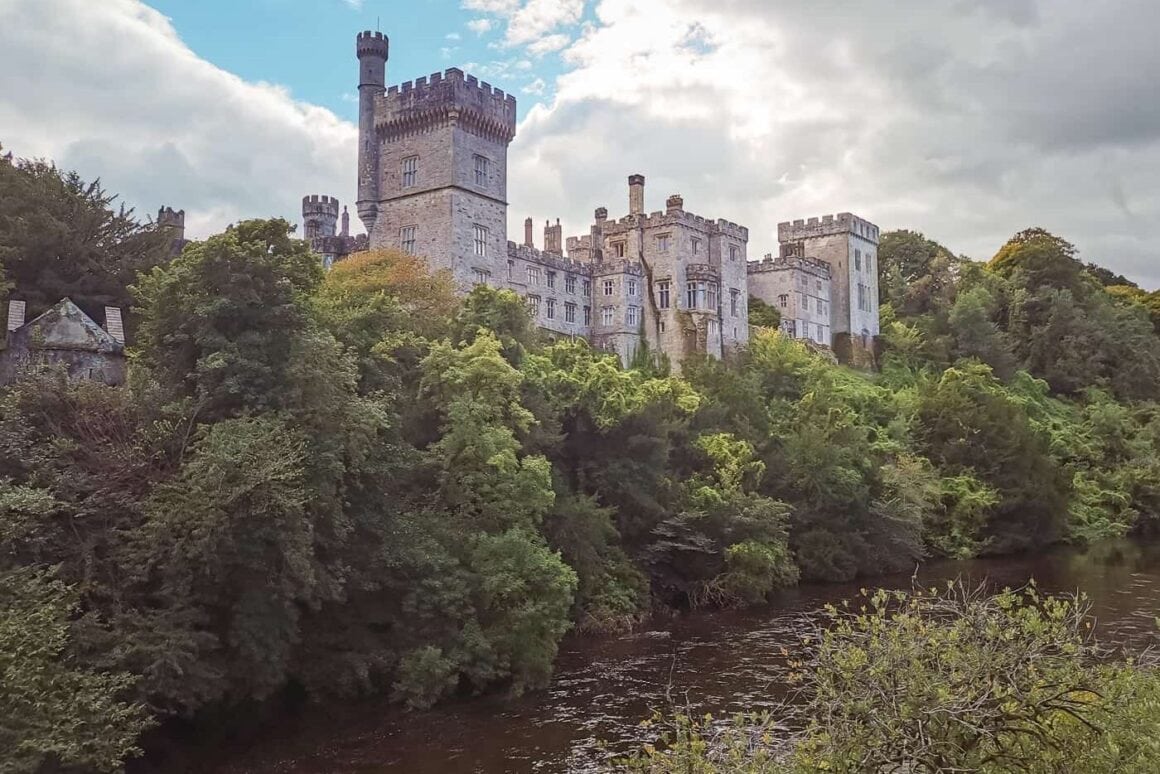 Large stone castle with towers and battlements rising above dense woodland beside a river in Ireland.