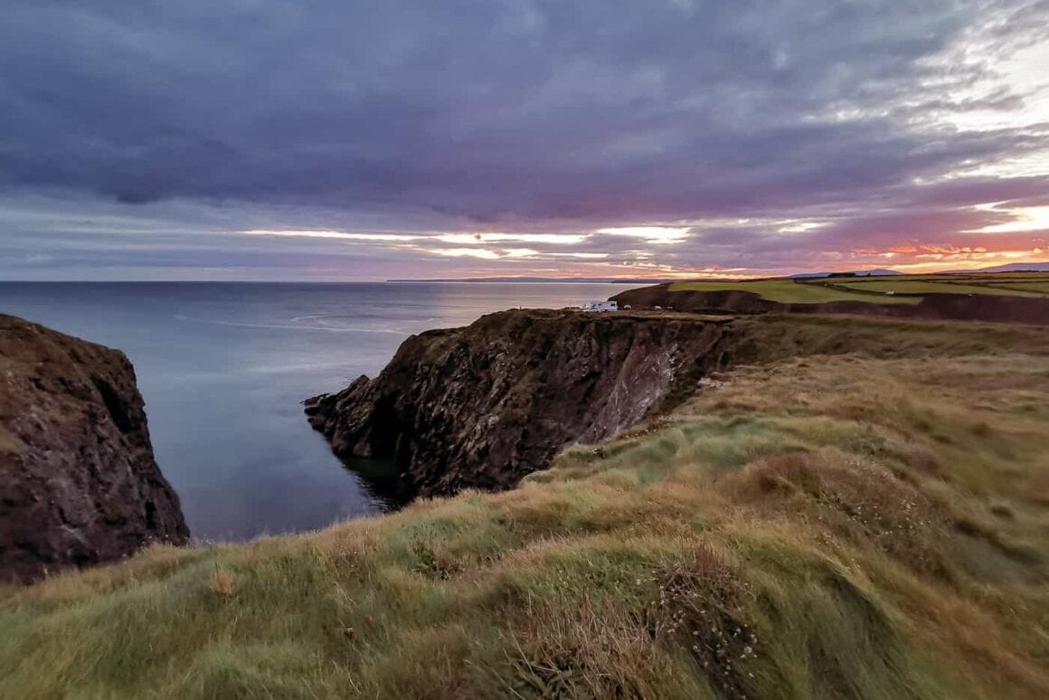 Dramatic rocky cliffs and grassy headlands overlooking the Atlantic Ocean beneath a colourful sunset sky in Ireland.
