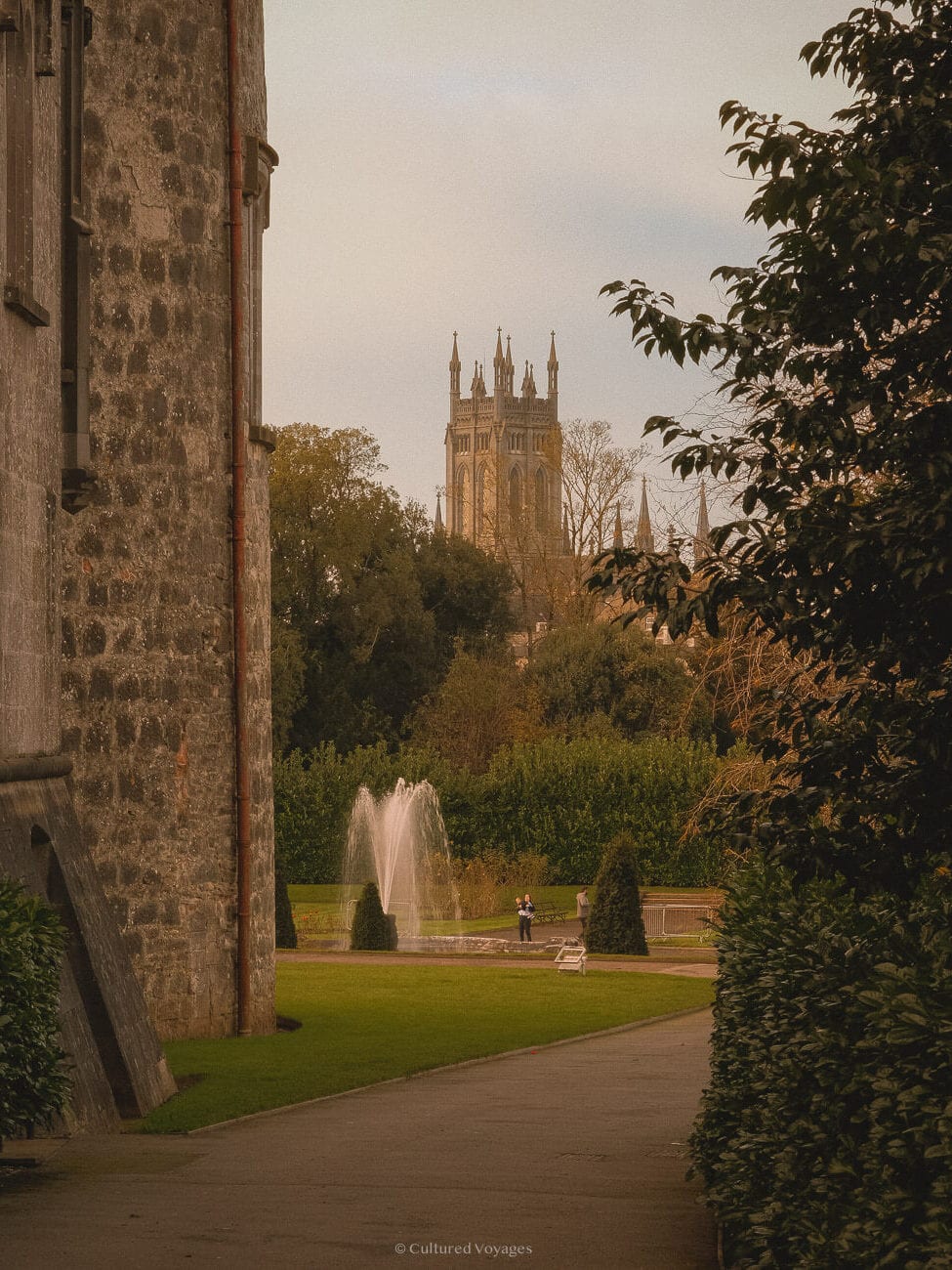A paved footpath alongside the medieval wall of a building, guiding towards a small park at the back with a fountain and a few people strolling. In the background, a cathedral tower emerges among the trees under clear blue skies in the Kilkenny Medieval Mile.