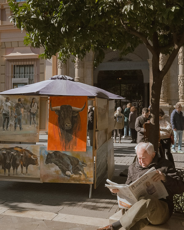 A man sitting in front of a tree with displays of paintings on one side under the shade of a tree while people walk in the background. Wondering how many days in Seville? Experience the art scene and leisurely strolls through the city streets.