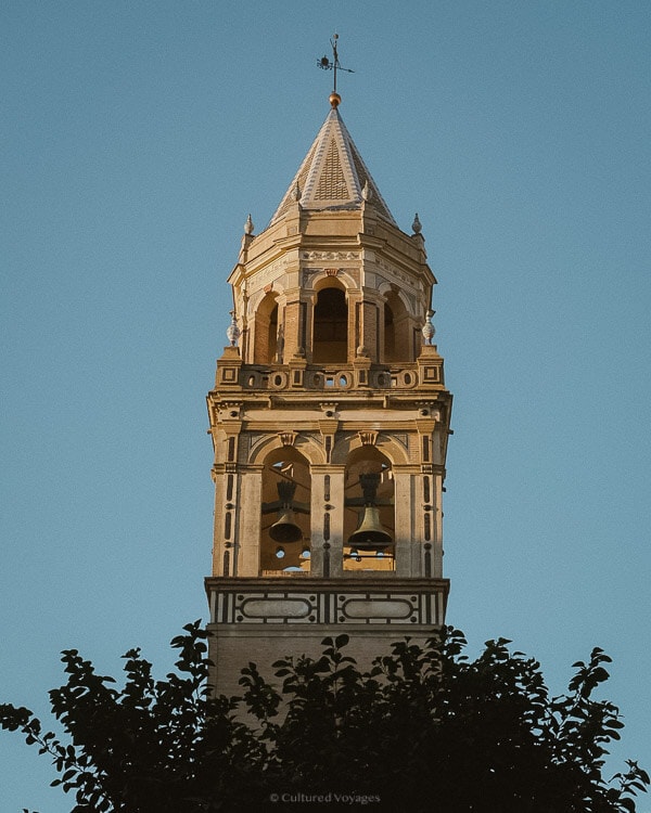 The bell tower of the Raimondello Orsini Tower featuring its cone roof with two bells under clear blue skies with some trees, while some trees can be seen in the foreground, evoking a serene atmosphere. Wondering how many days in Seville? Explore its beauty at your own pace.