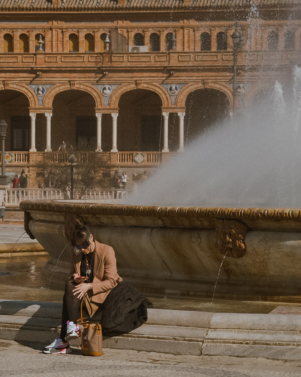 A person sitting on the side of a large fountain with the view of an old building behind it with large arched open walls. Wondering how many days in Seville? Enjoy serene moments by the fountain and explore the historic architecture of the city.
