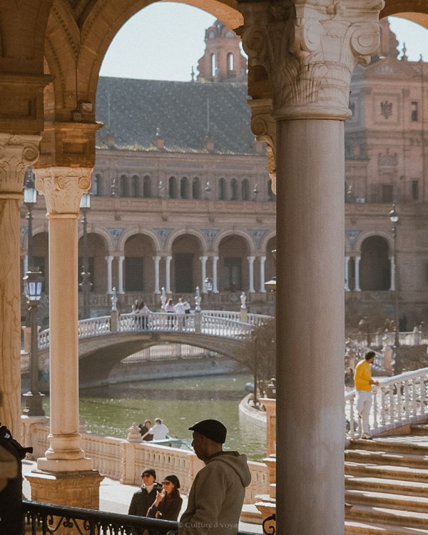 View of a river flowing under a bridge while people are walking and a view of a building can be seen in the back. Wondering how many days in Seville? Enjoy scenic walks along the riverbanks and explore the city's architectural beauty.