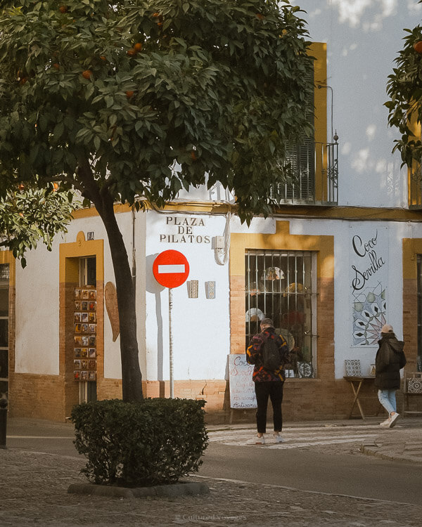 A street corner of a white walled building with few people walking while small trees can be seen in the foreground