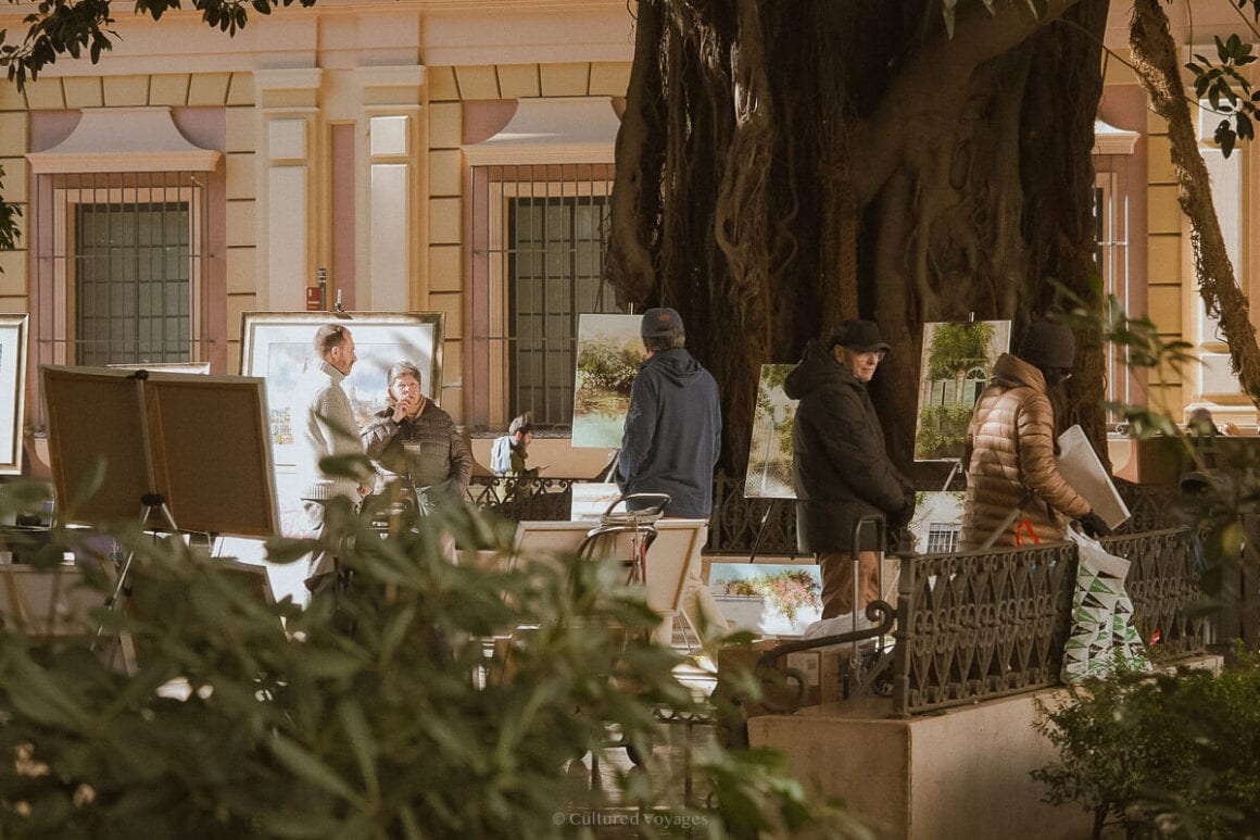 People looking at paintings under the shade of a large tree with a building in the background, while some leaves in the foreground. Wondering how many days in Seville? Delve into the art scene and cultural wonders of this city.