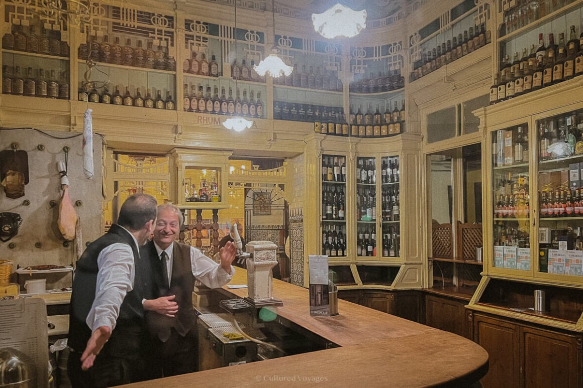 Two bartenders speaking with each other behind the bar surrounded by bottles of wine and liquor on the walls while lights drop down from the ceiling. Wondering how many days in Seville? Enjoy the lively atmosphere and delicious drinks in the heart of the city.