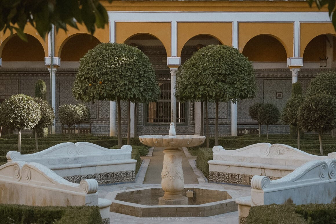 A small fountain surrounded by marble benches forming a circle in the middle of the garden with arched small trees in front of the building with arched open walls in yellow and white of the Casa de Pilatos. Wondering how many days in Seville? Immerse yourself in the architectural splendour and tranquil beauty of this historic city.