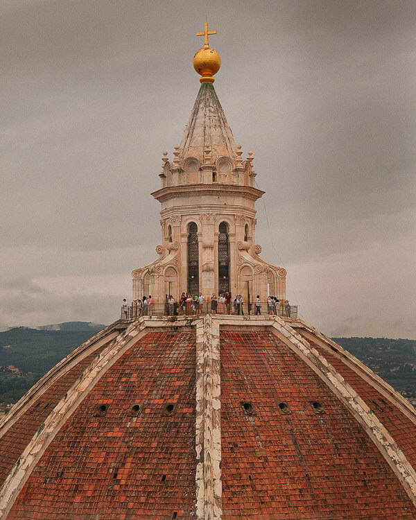 Top of the Florence Cathedral's dome with tourists at the observation deck, under a cloudy sky.