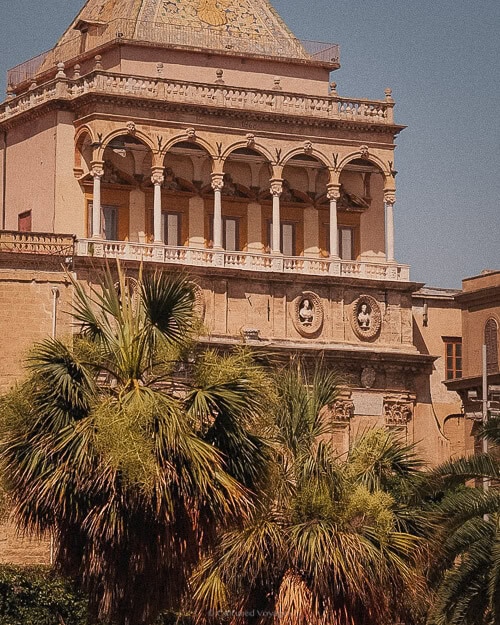 The exterior of a grand historic building in Palermo, Italy, featuring an ornate facade and lush palm trees in the foreground, representing the city's rich cultural heritage.