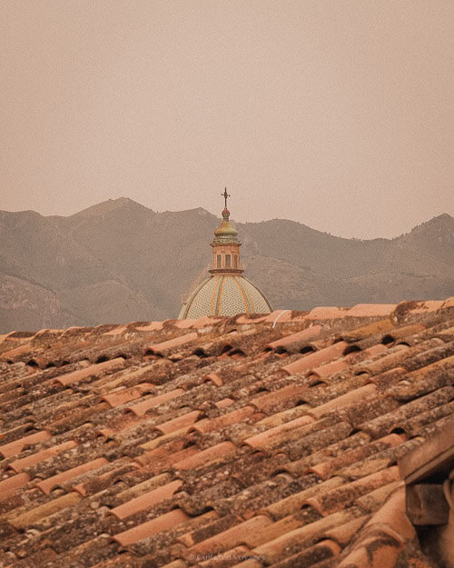 A distant view of a domed building peeking over terracotta rooftops in Palermo, with the mountains in the background, highlighting the beautiful and diverse landscape of the city.