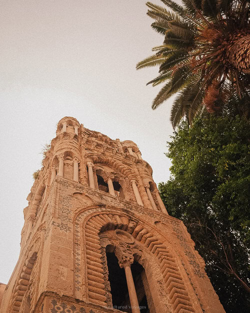 Looking up at an old stone tower in Palermo, surrounded by palm trees, its arches and carvings reflect a mix of architectural styles from different eras.