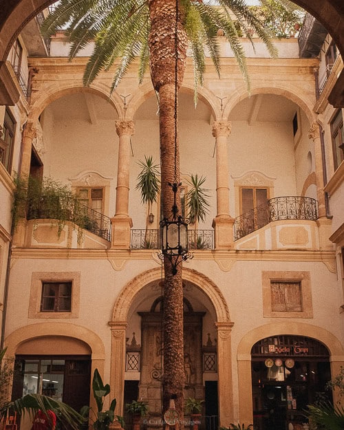 A grand courtyard in Palermo, featuring a tall palm tree in the centre, surrounded by arches and balconies, illustrating the city's blend of nature and classic architecture. Is Palermo worth visiting? This scene suggests it is.