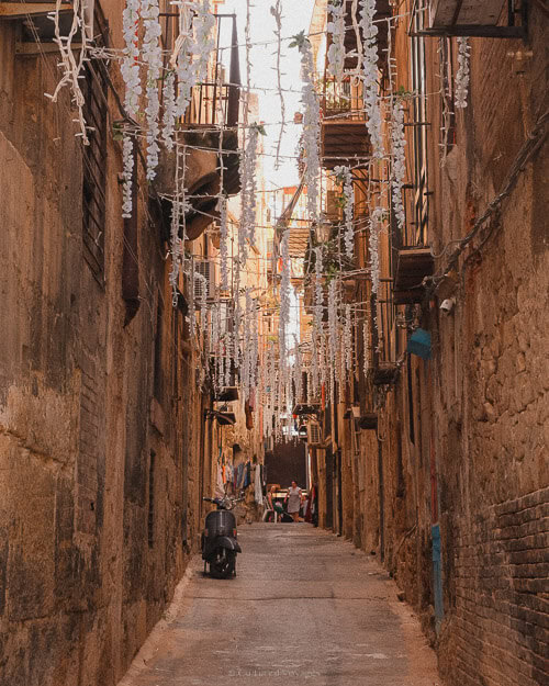 A narrow alleyway in Palermo, decorated with lights and lined with old, worn buildings, evoking the city’s vibrant and traditional atmosphere.