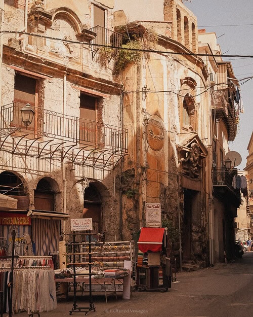 A bustling street market in Palermo, with colourful stalls selling fabrics and goods, set against the backdrop of an old, crumbling facade that speaks to the city's history.