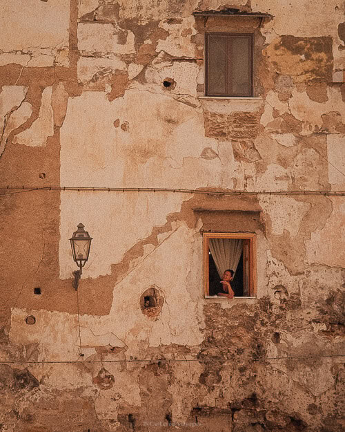 A weathered, old stone building with peeling plaster, showing a person peeking out from a small window while leaning on the sill.