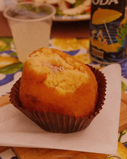 A close-up of a traditional Sicilian arancina, a golden fried rice ball with a meat filling, placed in a brown paper cup alongside a soft drink.