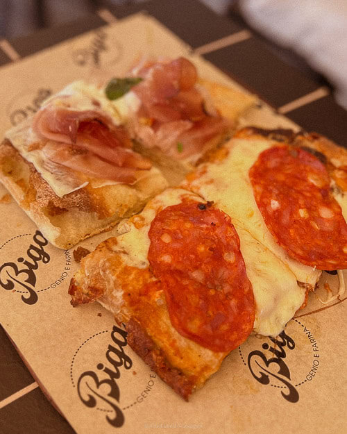Two slices of rectangular pizza with different toppings; one with cured meat and cheese, and the other with pepperoni, served on branded paper from Biga, an Italian pizzeria.