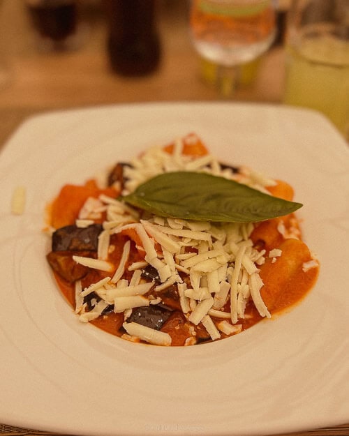 A plate of gnocchi topped with shredded cheese, aubergines, and a fresh basil leaf, served in a tomato-based sauce.