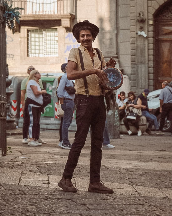 A street performer dressed in casual, vintage-inspired clothing, holding a tambourine with a star design. His wide smile and relaxed posture add to the lively, cultural atmosphere of the street, with onlookers and historic buildings in the background.