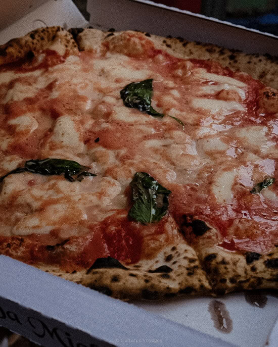 A Neapolitan pizza with melted cheese and tomato sauce, topped with fresh basil leaves. Served in a takeaway box, the pizza represents one of the authentic and delicious things to do in Naples for any food lover.