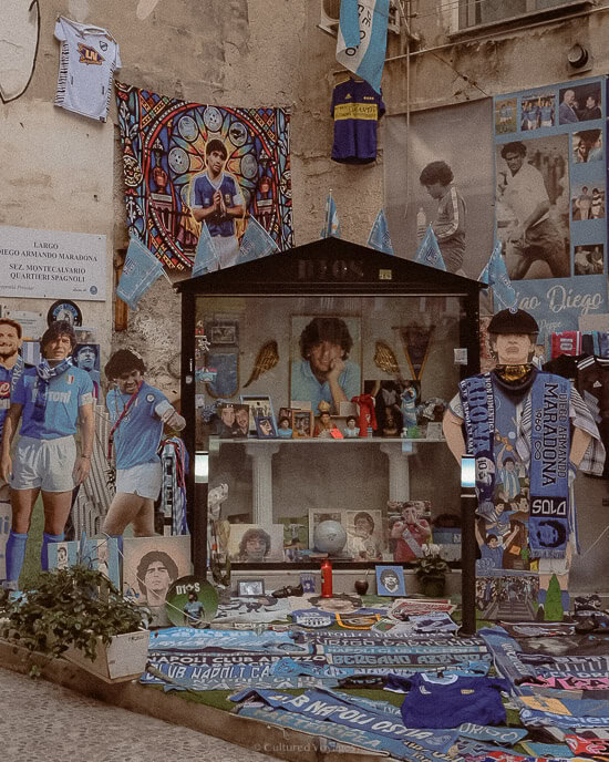 A small shrine dedicated to Diego Maradona, adorned with photographs, scarves, and memorabilia celebrating the legendary footballer. Flags, posters, and souvenirs cover the walls, reflecting the deep admiration and love for Maradona in Naples.