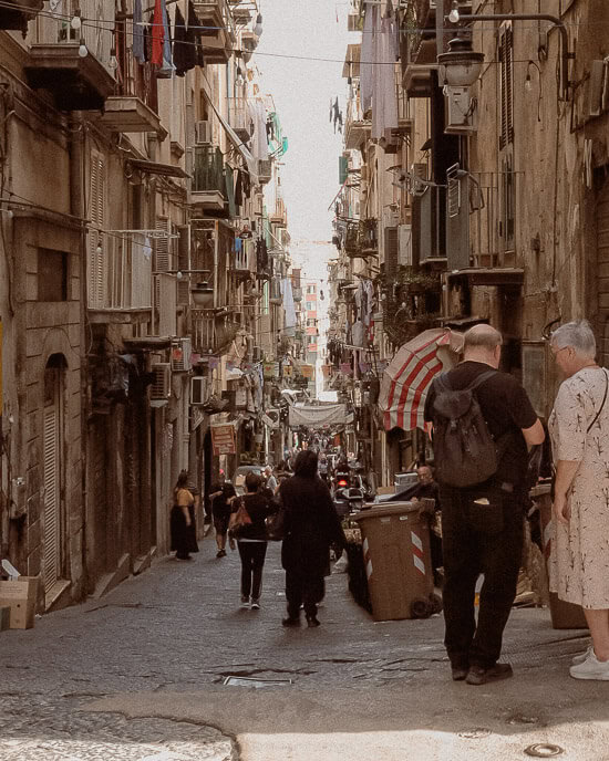 An inclined street in Naples bustling with people walking past small shops and residential buildings. The narrow, lively street scene captures everyday life in a historic Italian neighbourhood, where locals and tourists alike explore the city.