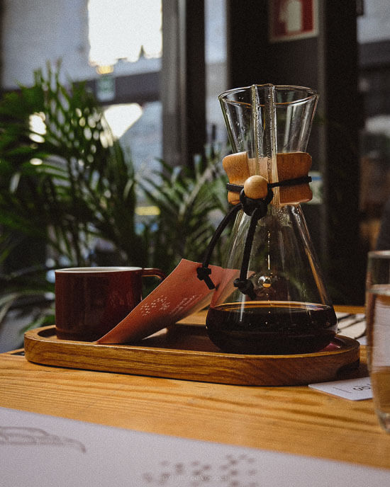 A wooden tray holding a Chemex coffee maker and a red mug, sitting on a sunlit table inside a cozy café in Porto.