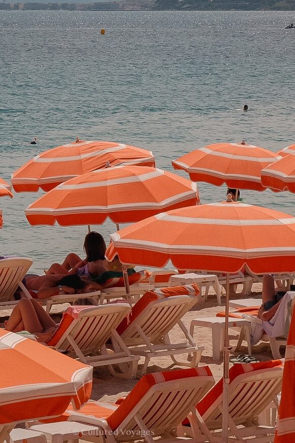 Beachside packed with people on orange and white sunlounges under orange and white striped umbrellas with a view of crystal blue waters in the background with people swimming - visiting antibes guide