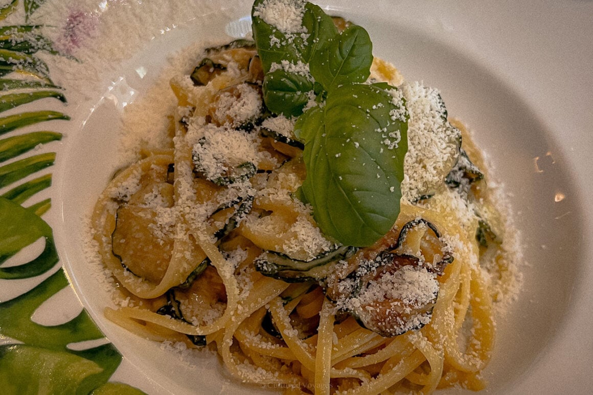 A close-up of a plate of spaghetti alla Nerano, topped with thinly sliced zucchini, freshly grated Parmesan cheese, and a sprig of basil, served on a white plate with a green leaf design.