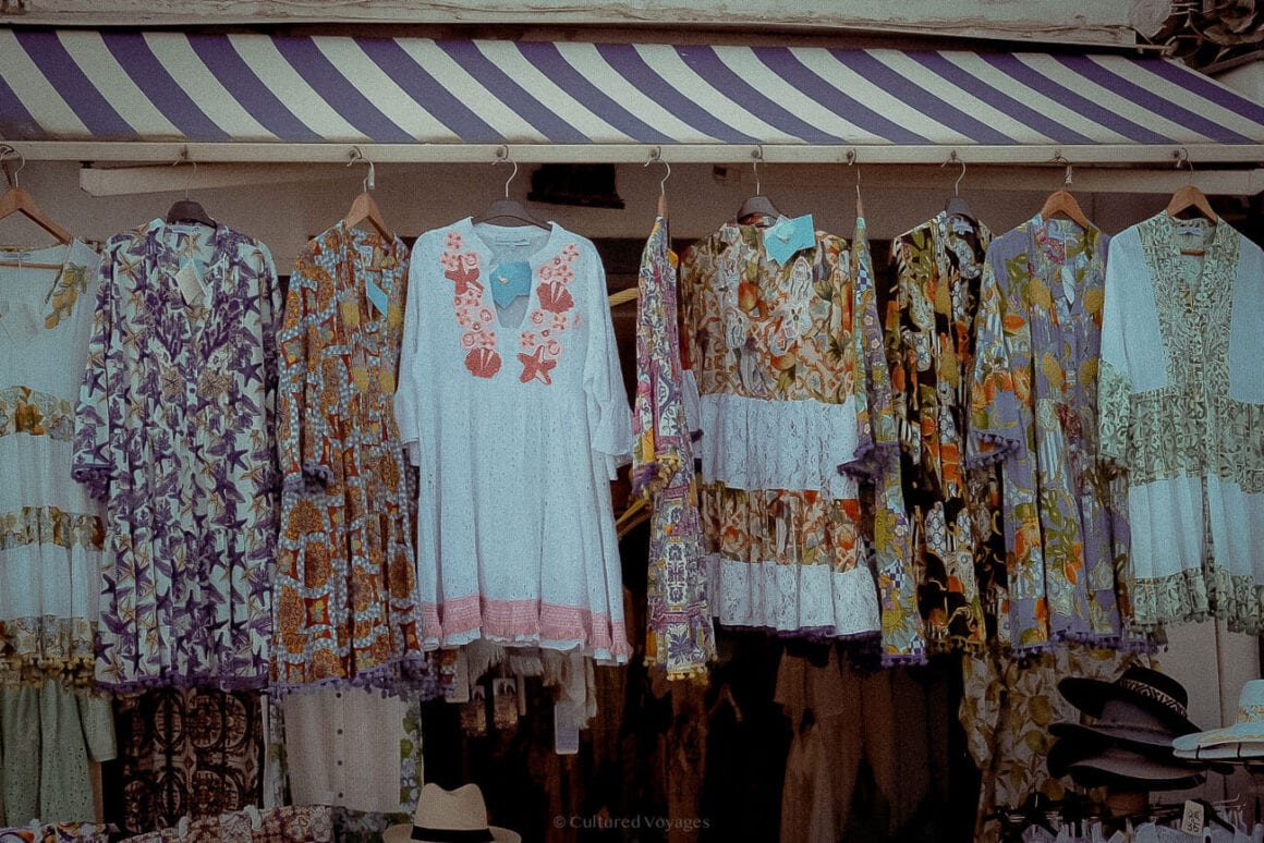 A colourful display of bohemian-style dresses hanging on wooden hangers under a striped purple-and-white awning at a market stall.