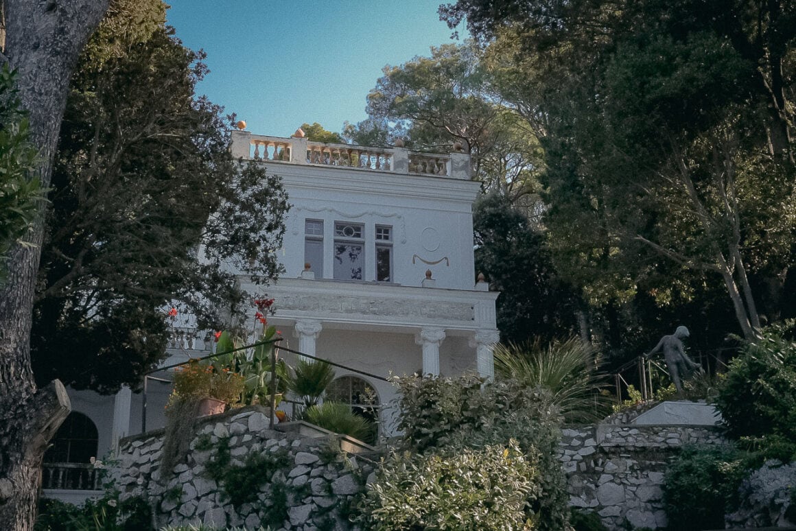 A grand white villa with ornate details, surrounded by tall trees and vibrant greenery, with a bronze statue of a dancing figure in the garden.