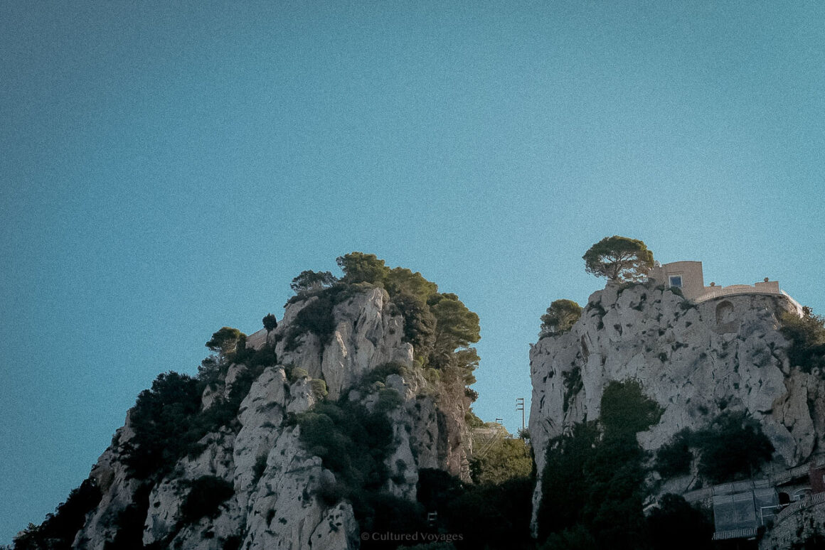 Two rugged limestone cliffs topped with pine trees and a yellow villa, standing prominently against a bright blue sky.