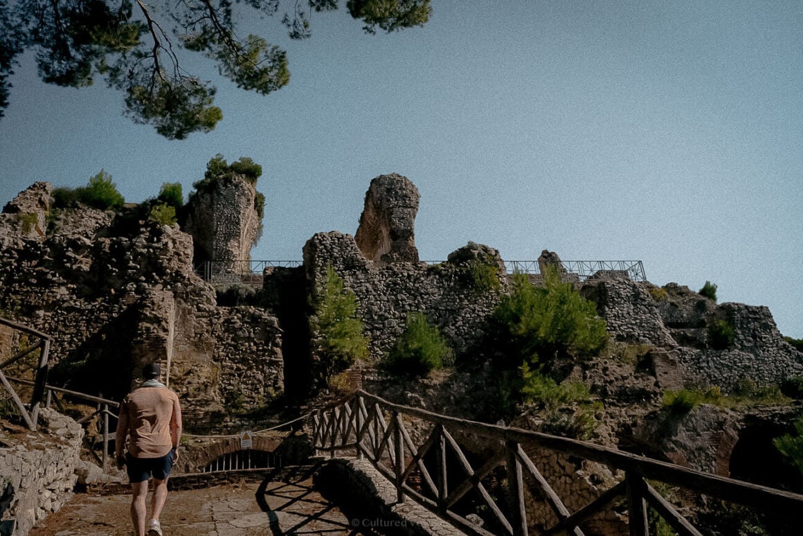 A man walking along a wooden path leading to the ruins of a historic stone structure, surrounded by scattered greenery under a clear sky.