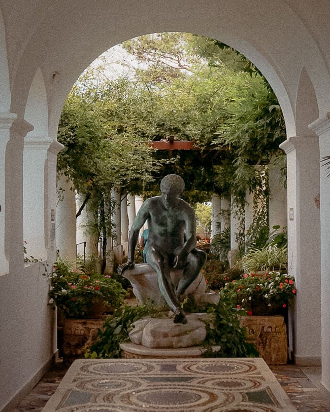 A bronze statue of a seated figure, surrounded by greenery and colourful potted flowers, placed at the centre of an arched courtyard.
