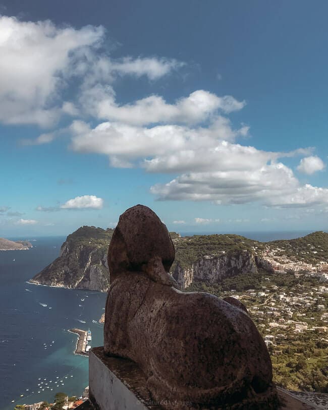 A breathtaking view of Capri's coastline, with a weathered stone sphinx statue overlooking a sweeping expanse of sea, cliffs, and villas, captured during a Naples to Capri Day Trip.