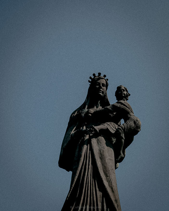 A statue of the Virgin Mary holding a child, standing against a clear blue sky, showcasing detailed craftsmanship and a serene expression.