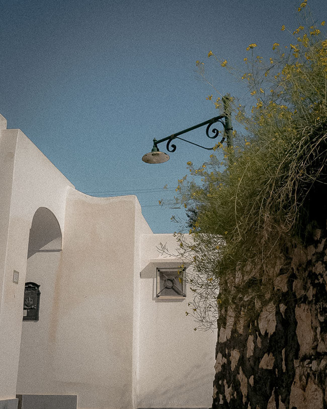 A minimalistic Mediterranean building with white walls, a black lamp, and yellow wildflowers growing from a stone wall under a bright blue sky.