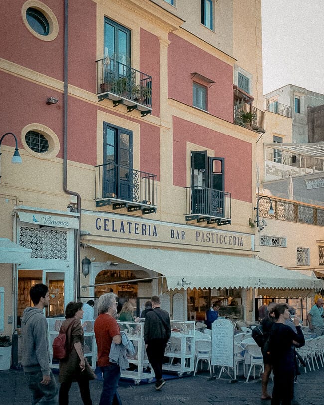 A bustling gelateria and pasticceria with a red-and-yellow facade, featuring outdoor seating and people enjoying the lively atmosphere.