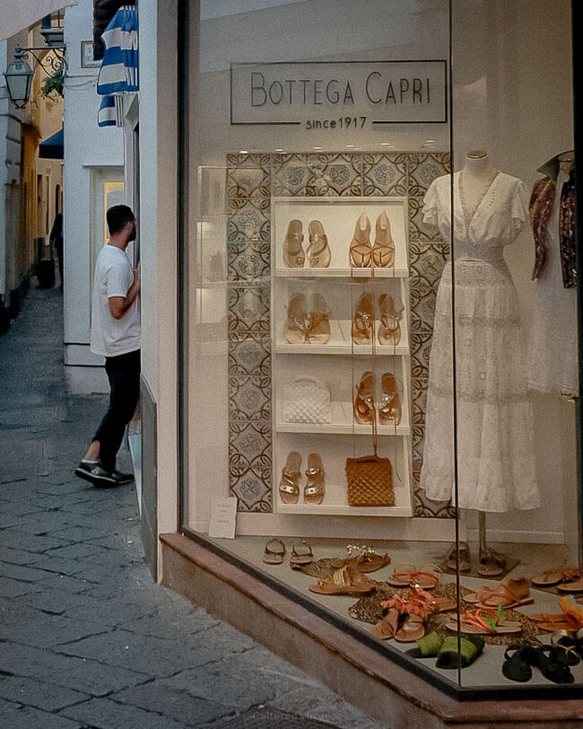 A shop window of "Bottega Capri," showcasing handcrafted leather sandals and a white lace dress, set against patterned tiles, with a man standing near the entrance in a narrow cobbled alley.