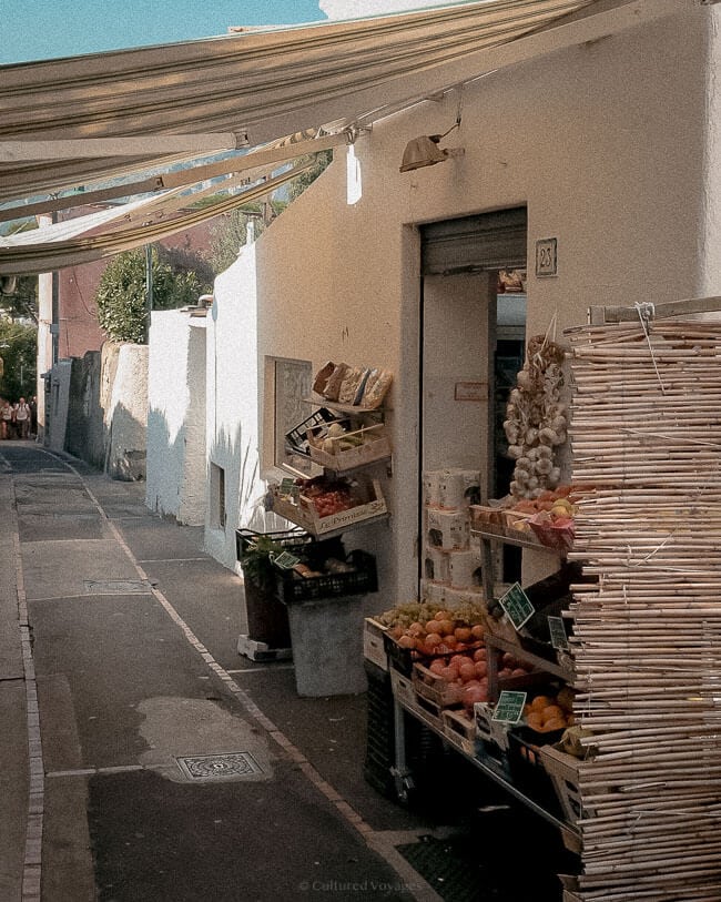 A small market stall along a narrow street, displaying fresh produce, garlic strings, and packaged goods under a striped canopy, with sunlight casting soft shadows on the whitewashed walls.