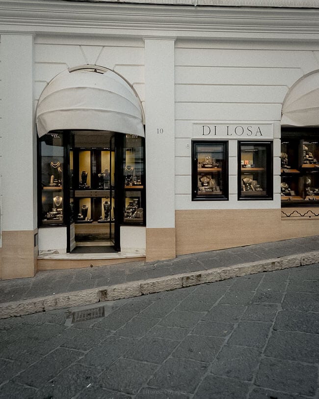The storefront of a jewellery shop named "Di Losa," featuring elegant displays of gold and silver jewellery in a minimalist white and beige exterior, with arched windows and a stone pavement.