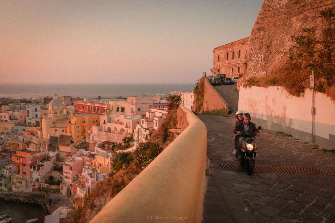 A couple riding a scooter along a narrow, winding street overlooking Procidaโs iconic multicoloured buildings and the coastline, framed by warm evening light.